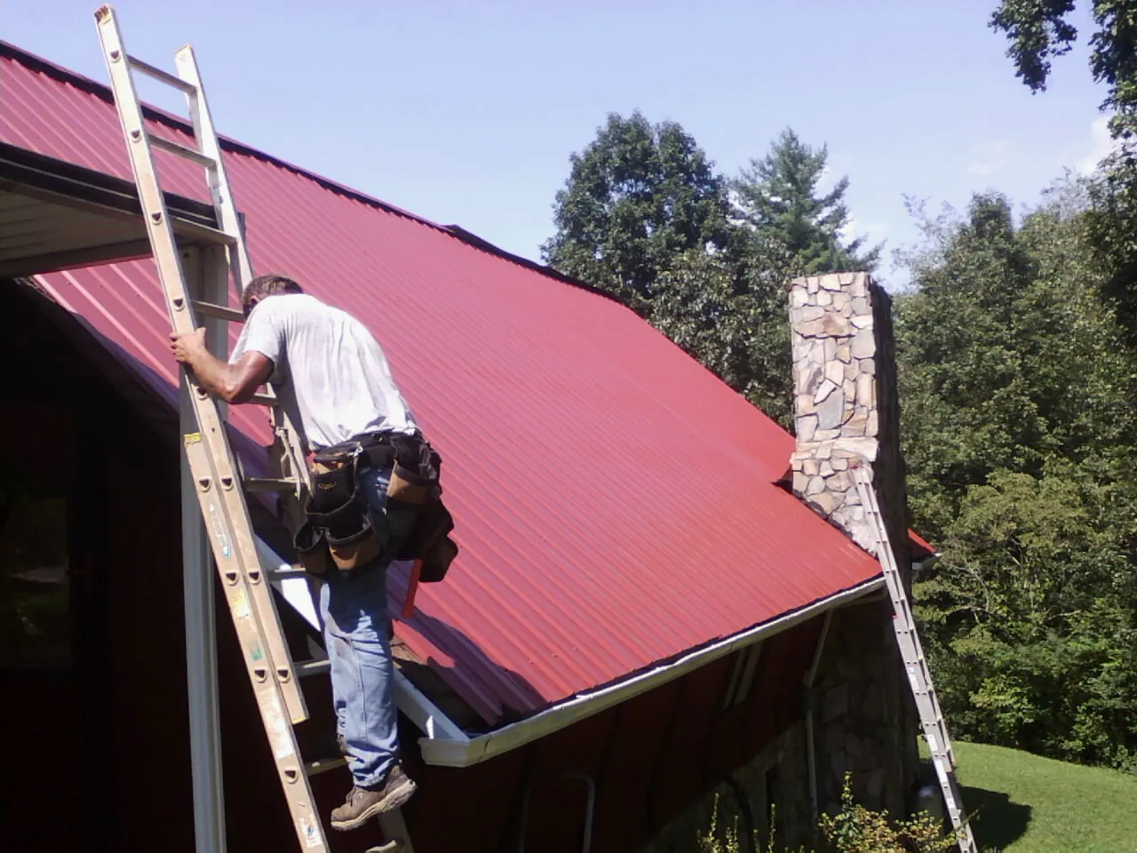 Roofer on a ladder beside a red metal roof and stone chimney — Soffit & Fascia Repair by Roofing Brothers in Jakin